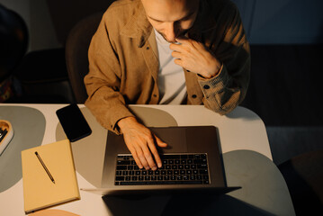 A man works at night on a laptop computer at home under the light of a desk lamp, worried and feeling exhausted