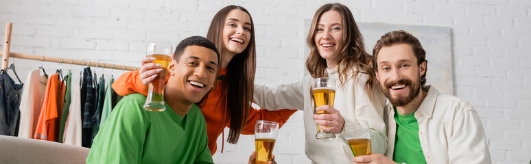 cheerful women hugging with interracial men holding glasses of beer in living room, banner