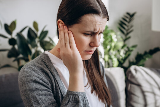 Close Up Portrait Of Upset Young Girl Holding Painful Ear, Suddenly Feeling Strong Ache. Unhealthy Caucasian Woman 20s Suffering From Painful Otitis Sitting On Sofa At Home. Health Problems Concept