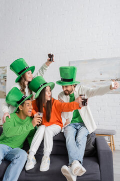 cheerful bearded man taking selfie with happy interracial friends holding glasses of dark beer on Saint Patrick Day