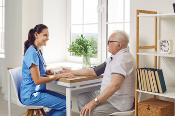 Smiling female doctor measuring pressure of elderly patient. Side view portrait of senior man and young nurse sitting at table and communicating in clinic. Healthcare and medicine concept