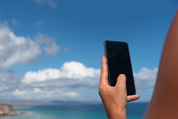 Primer plano del brazo de un hombre bronceado mirando su tel&eacute;fono m&oacute;vil en el fondo de la playa de arena blanca y el mar turquesa de Jandia, en un d&iacute;a soleado en Fuerteventura, Islas Canarias.