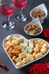 Varieties of elite cheese in heart-shaped plate, cashews, peanuts and two glasses of wine for Valentine's Day on gray background
