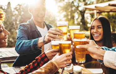 Happy friends cheering beer glasses at brewery pub garden - Group of young people enjoying happy hour sitting at bar table - Life style concept with guys and girls having dinner party together - obrazy, fototapety, plakaty