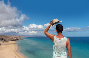 Hombre con camiseta verde quitándose el sombrero de paja frente a la playa de arena blanca y el...
