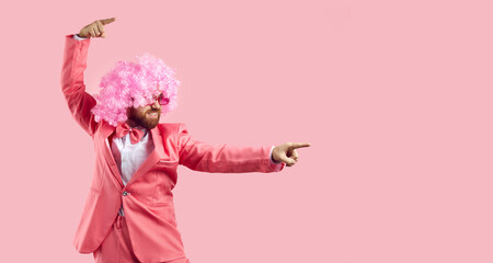 Side view of happy funny man with ginger beard, in suit, bow tie and curly clown wig standing on pink background invites to fun party, points index fingers away and shows something on blank copyspace