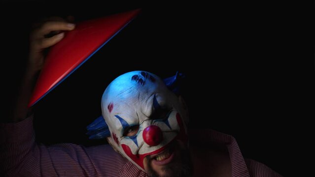 A scary clown in a red striped shirt is holding a red lampshade with a blue lamp in his hand above his head. A bearded man in an evil clone mask is grimacing and fooling around looking into camera