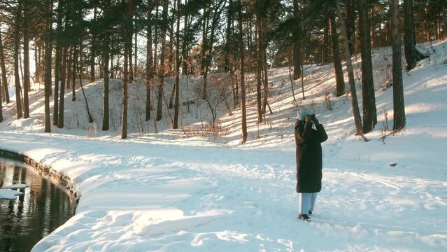 Beautiful young female photographer in blue hat, eyeglasses and black jacket walking and taking pictures on photo camera at winter time in the park covered snow at sunny day. Slowmotion.