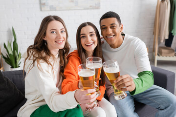 cheerful women and african american man clinking glasses of beer while looking at camera in living room