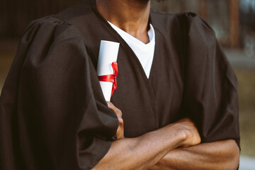 Portrait closeup of black man graduate from university stand outdoors in mantle with crossed arms and holding higher education diploma, selective focus. Student, graduation, ceremonial celebration.