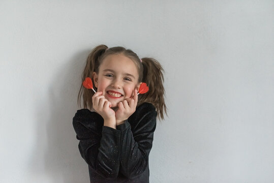 pretty and smiling seven year old girl with two ponytails wearing black velvet bodysuit is holding two red heart shaped lollipops near her face while standing on white background