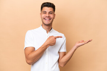 Young caucasian man isolated on beige background holding copyspace imaginary on the palm to insert...