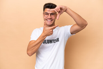 Young volunteer caucasian man isolated on beige background focusing face. Framing symbol