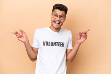 Young volunteer caucasian man isolated on beige background pointing finger to the laterals and happy