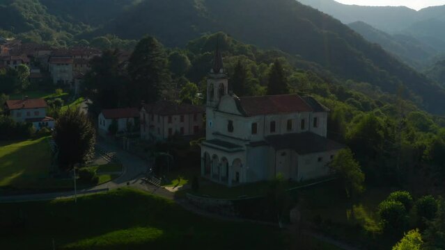 Aerial View Of Scenic Italian Village Parish Church Located Beside Forested Hills In Arola, Italy. Dolly Forward, Tilt Up