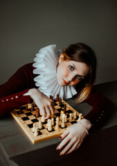 A beautiful woman in burgundy clothes and a white jabot sits at a table and plays wooden chess. The Queen's gambit. Mind and logic. Jester. - obrazy, fototapety, plakaty