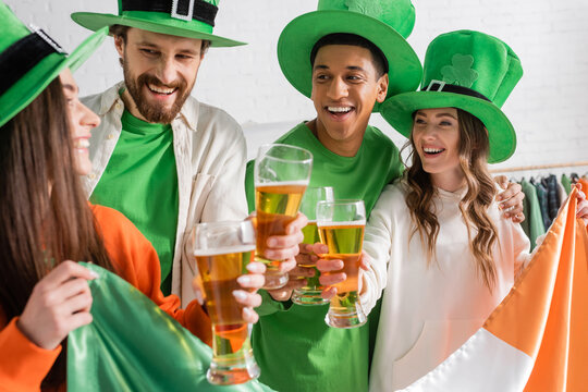 happy and interracial friends in green hats holding glasses of beer and Irish flag while celebrating Saint Patrick Day - Powered by Adobe