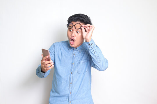 Portrait of shocked Asian man in blue shirt looking at mobile phone. Wow face expression. Isolated image on white background