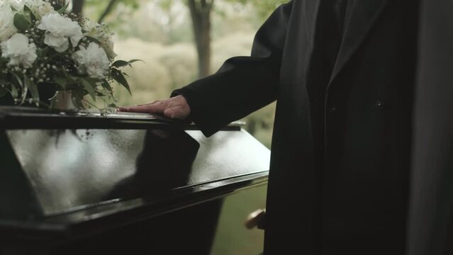 Cropped shot of unrecognizable man in long black coat putting his hand on black wooden lid of coffin at funeral outdoors