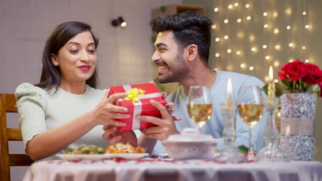 excited man giving Gift to girlfriend during candlelight dinner at home - concept of wedding Anniversary or birthday present, romantic night and valentines day.