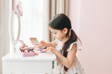 a beautiful little girl in the mirror preening. a little girl is sitting at a children's table and is wearing children's cosmetics.