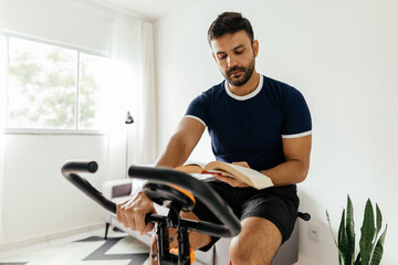 Man working out on exercise bike while reading a book at home