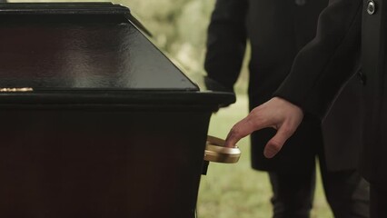 Cropped shot of unrecognizable pallbearers carrying elegant black wooden coffin along cemetery during funeral ceremony