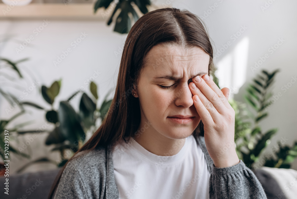 Wall mural Close up of sad woman suffering from strong eye pain. Eyesight problem. Tired young brunette female touching eyes sitting on sofa at home interior living room. Injuries Poor health Illness concept