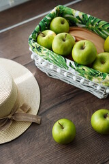 A top view of a white wicker basket with green juicy apples on a wooden table near the window. Next to the basket lies a beige hat and a walnut basket. Sunny day.  Copy space