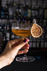 Cocktail Angel Face fatto con apricot brandy, calvados e gin arte, guarnito con arancia essiccata mentre viene tenuto in mano da una cliente di un cocktail Bar serale
