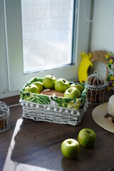 A white wicker basket with green juicy apples on a wooden table near the window. Next to the basket lies a beige hat and a walnut basket. Sunny day. Beautiful background