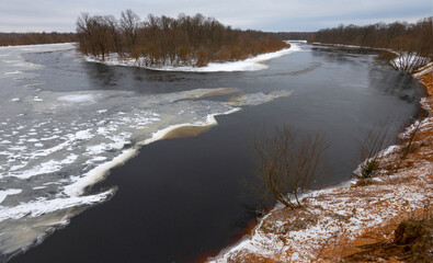 Winter landscape with a partially frozen river. the flow of water. Islet and birch grove. Steep steep bank. Panorama