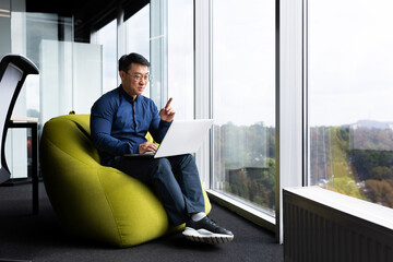 Video call, online remote conversation with clients and colleagues, successful asian man sitting in office near window on soft chair and talking remotely in meeting, man smiling using laptop at work. - Powered by Adobe