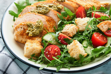 Fresh salad with grilled chicken fillet, arugula, tomatoes, cucumber and pesto sauce, sesame seeds with olive oil in white bowl on light slate background. Healthy lunch menu. Diet food. Top view.