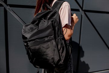 The cropped photo of the bagpack. The modern girl teenager dressed black t-shirt, jeans, sunglasses, watches and bagback walking in the street at the background grey wall