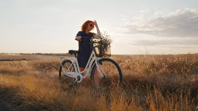 Happy Woman walks through wheat field enjoying with bicycle bouquet of field daisies in sunset sunlight in summer. Go Everywhere. Rest from city life. Reloading people among nature. Relax. Travel