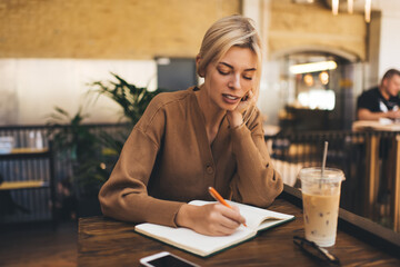 Caucasian female student making informative notes during coffee time in cafe interior, young blonde woman with education notebook for learning writing text publication while doing homework indoors