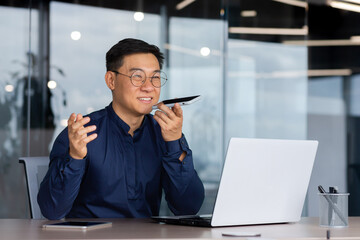 Successful asian man inside office working with laptop, businessman recording audio message using app on smartphone, man using artificial intelligence assistant to help finding a solution.