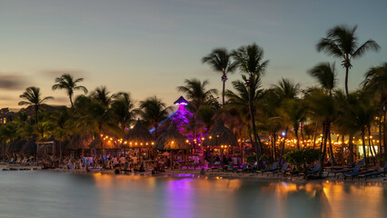 A tastefully lit beach bar, on a tropical beach next to the shore.  The colourful lights are reflecting in the sea.