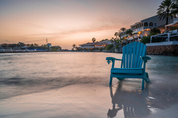 An Adirondack chair, on the shoreline of a tropical beach, at sunset.  The ocean is lapping at the chair. 