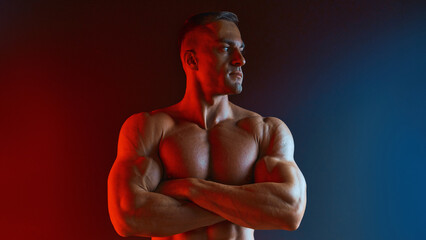 Single standing young handsome serious male bodybuilder on red-blue creative light