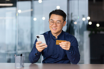 Satisfied and smiling man inside office at work, using app on phone and bank credit card for online...