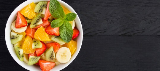 Fruit salad in the white bowl on the black wooden background. Top view. Copy space. Close-up.