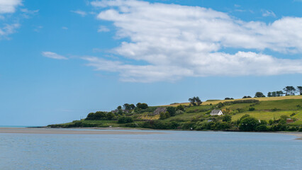 Beautiful sky over a calm water surface on a summer day. The picturesque green coast of Ireland. Several buildings on the hill. Seascape. Green trees on island