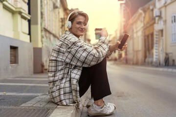 Fototapeta na wymiar Stylish young woman in headphones sitting on curbside in city holding disposable coffee cup in hand looking at camera and smiling.