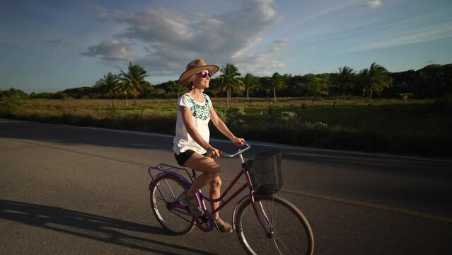 Mature senior woman holding her hat while biking into the sun wearing sunglasses in a tropical setting. Filmed in slow motion.