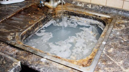 Tap water flows into a dirty kitchen sink. Dirty soapy water for washing dishes in the sink on the kitchen table. House cleaning. Water faucet with flowing water. Washing dishes from food leftovers