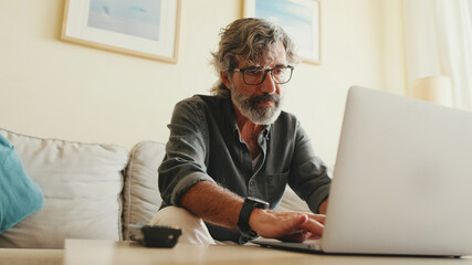 Elderly positive man typing on laptop sitting on sofa in living room