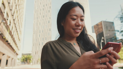 Fototapeta premium Close-up of young happy businesswoman using mobile phone while standing on modern buildings background