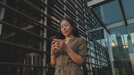 Young happy businesswoman using mobile phone while standing on modern buildings backgroun